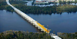 An aerial view of the US-17 Washington Bypass, constructed by the United Infrastructure Group, spanning over a serene river. Dense trees flank both sides of the river, and a cluster of buildings can be seen in the distance. A large yellow crane is positioned on the bridge, indicating ongoing work.