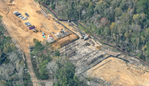 Aerial view of Berlin G Myers Parkway Phase III construction site overseen by United Infrastructure Group, showcasing vehicles, equipment, and structural developments amidst a wooded area.